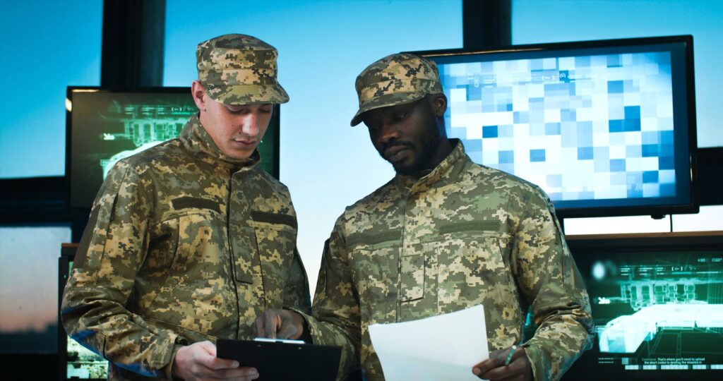 Two uniformed military officers reviewing documents in a command center in front of monitor screens.