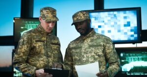 Two uniformed military officers reviewing documents in a command center in front of monitor screens.