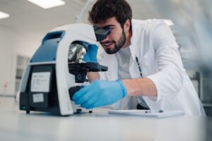 A scientist in a lab coat and gloves examining a sample through a microscope in a research laboratory.