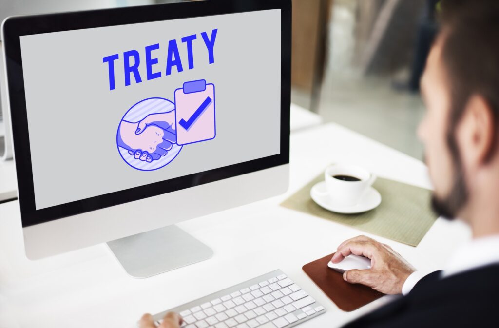 A person at a desk reviewing a treaty agreement displayed on a computer screen, with a handshake and checkmark icon.