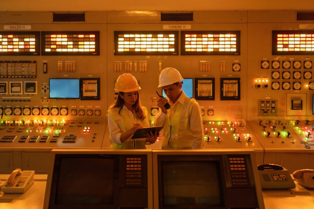 Two plant operators in hard hats reviewing controls and switches inside a power plant control room.